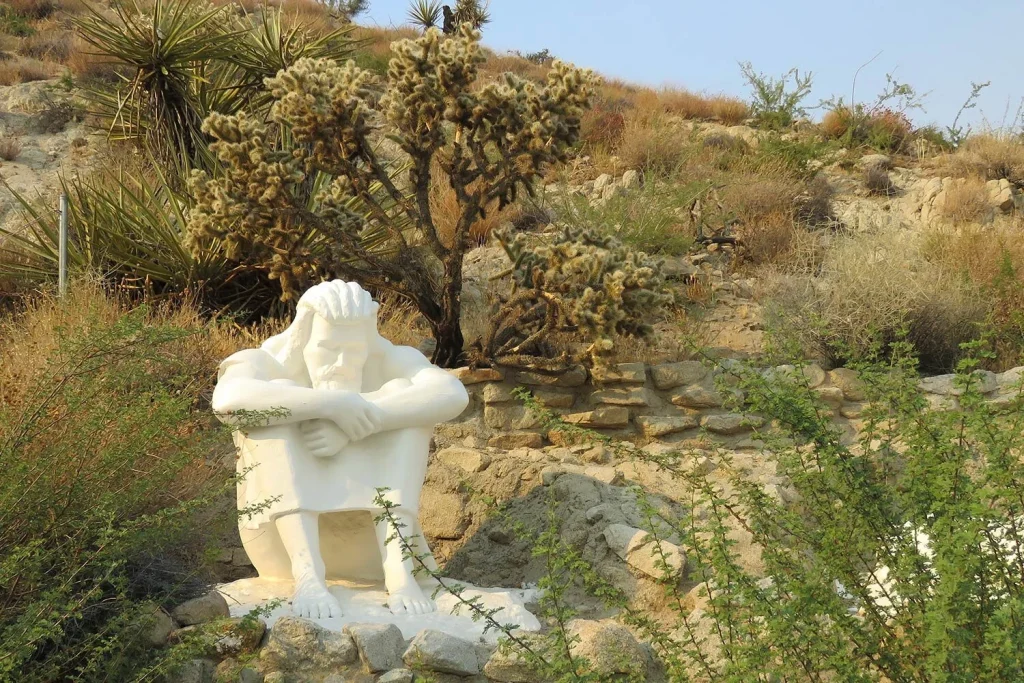 A white statue of Jesus sitting with arms on knees in a desert garden